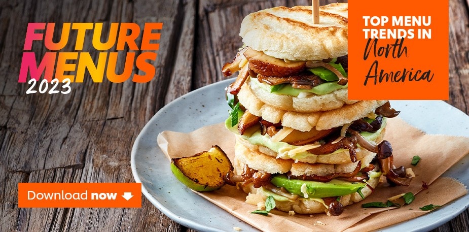 A tall, professional plated sandwich is shown on a wooden table. The text reads: Future Menus 2023. Top Menu Trends in North America. Download Now.